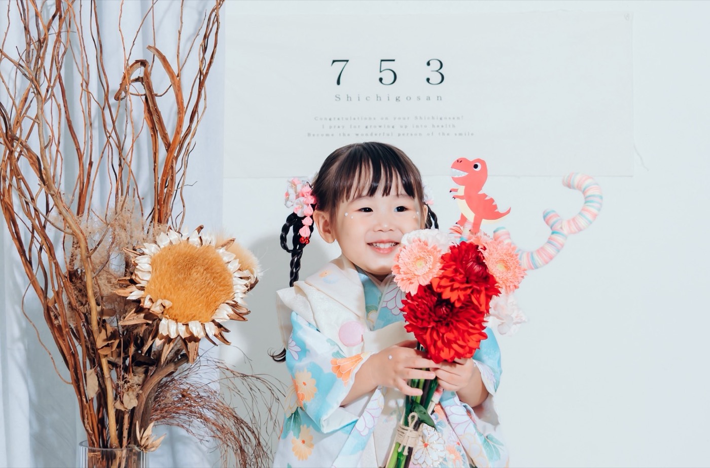 七五三の着物姿のお子さんが、赤いダリアとサーモンピンクのガーベラの花束を持って笑顔|フローリスト・つつみ